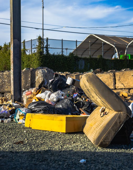 A pile of mixed rubbish including several black plastic trash bags, a yellow plastic storage container, and an old, worn car seat cushion with fabric upholstery, all situated on a gravel surface in an