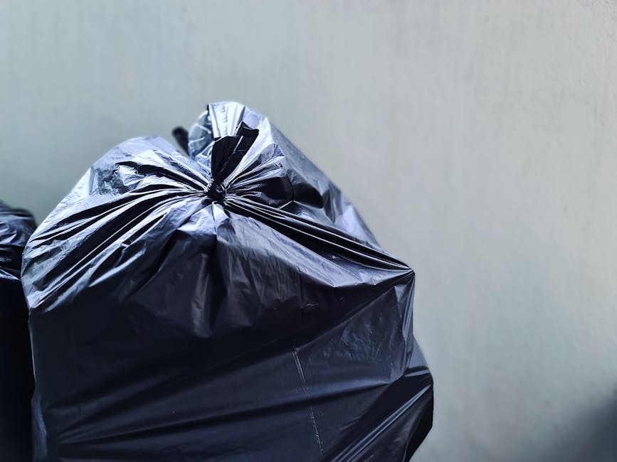 A pile of mixed rubbish including several black plastic trash bags, a yellow plastic storage container, and an old, worn car seat cushion with fabric upholstery, all situated on a gravel surface in an