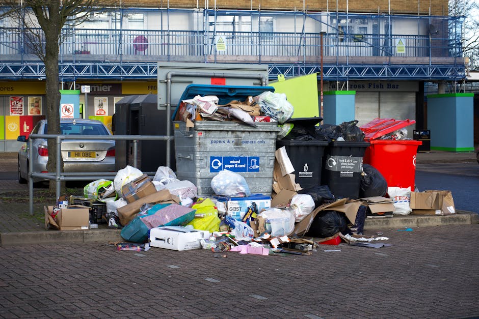 A collection of mixed household waste and rubbish bags piled on a gravel surface in an outdoor environment, with a weathered, beige car seat tipped over in the foreground. The waste appears to include