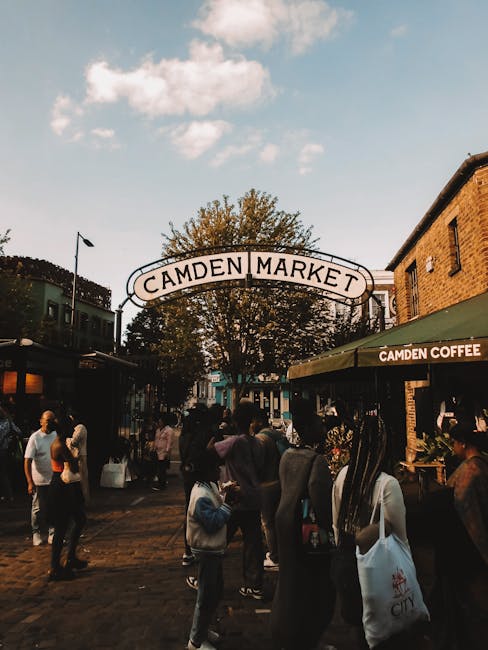 A busy street scene at Camden Market with a variety of pedestrians walking along the pavement, some carrying shopping bags or pushing strollers. In the background, there is a prominent arched sign tha